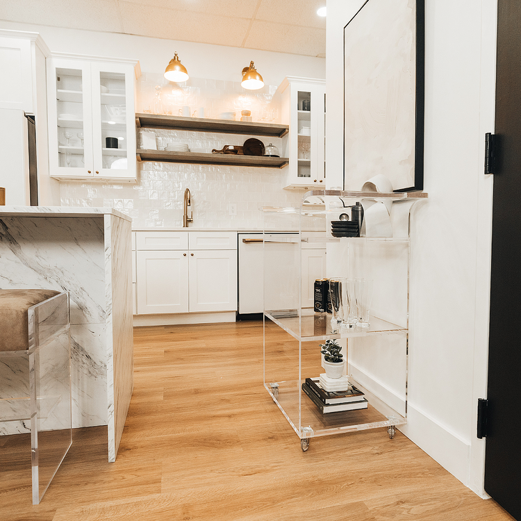 A clear acrylic bar cart featuring 3 shelves, wheels, and clear acrylic handles decorated with bar essentials, candles, and other decor items placed against a kitchen wall.