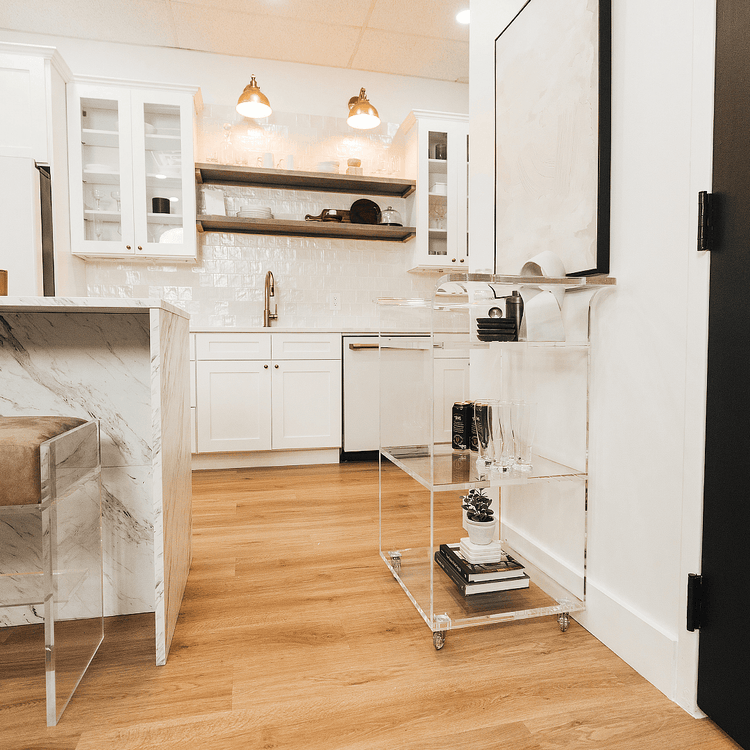 A clear acrylic bar cart featuring 3 shelves, wheels, and clear acrylic handles decorated with bar essentials, candles, and other decor items placed against a kitchen wall.