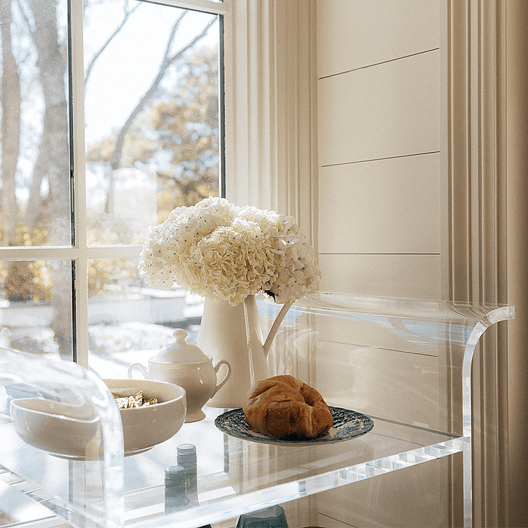 A clear acrylic bar cart with flowers, drinks, and snacks placed in front of a window in a living room setting.