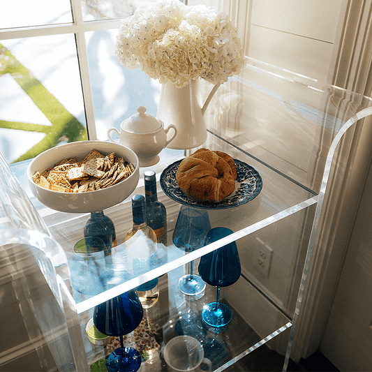 A clear acrylic bar cart with flowers, drinks, and snacks placed in front of a window in a living room setting.