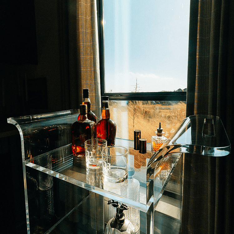 A clear acrylic bar cart with spirit bottles and glasses placed in front of a window in a darkened living room setting with reflections of the sunlight outside shining through the window.