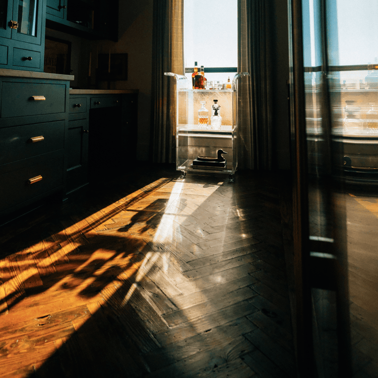 A clear acrylic bar cart with spirit bottles and glasses placed in front of a window in a darkened living room setting with reflections of the sunlight outside shining through the window.