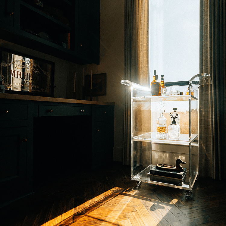 A clear acrylic bar cart with spirit bottles and glasses placed in front of a window in a darkened living room setting with reflections of the sunlight outside shining through the window.