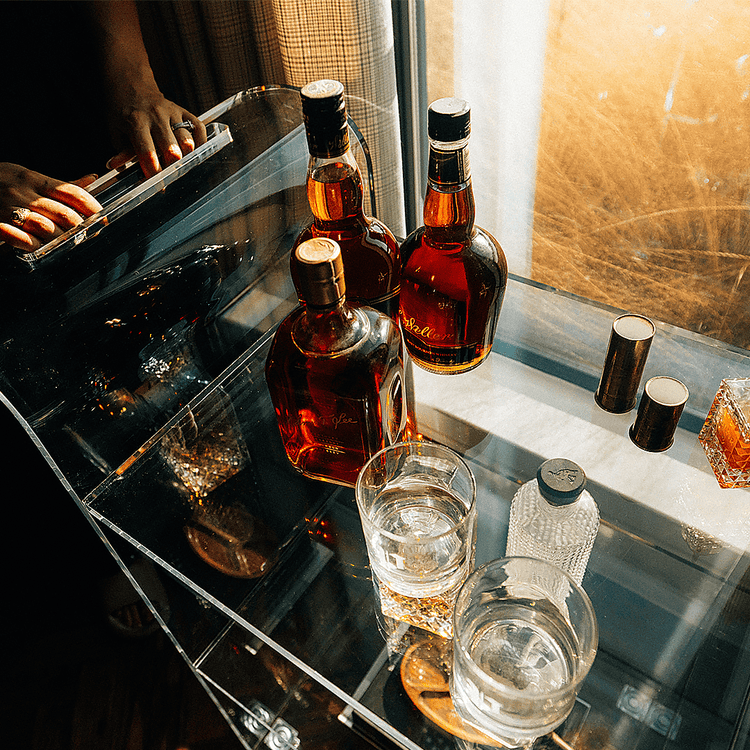 A clear acrylic bar cart with spirit bottles and glasses placed in front of a window in a darkened living room setting with reflections of the sunlight outside shining through the window.