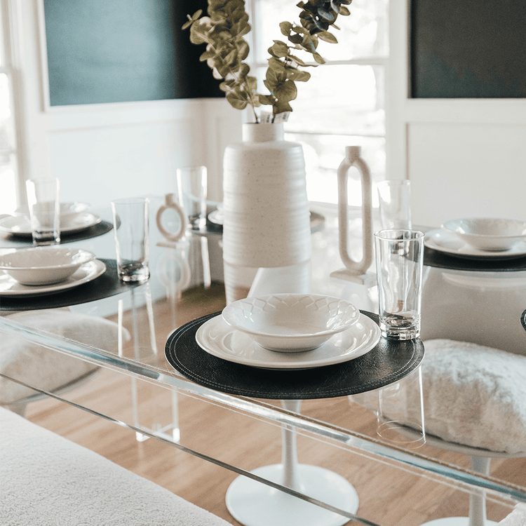 A clear acrylic lucite dining table decorated with flowers and dinner place settings surrounded by white modern chairs and a white padded bench for seating in a brightly lit dining room.