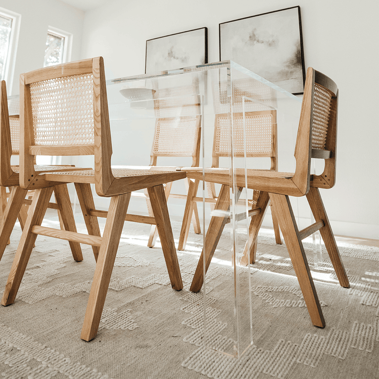 A clear acrylic lucite dining table decorated with a bowl of fruit surrounded by light wood and wicker chairs for seating in a brightly lit dining room.