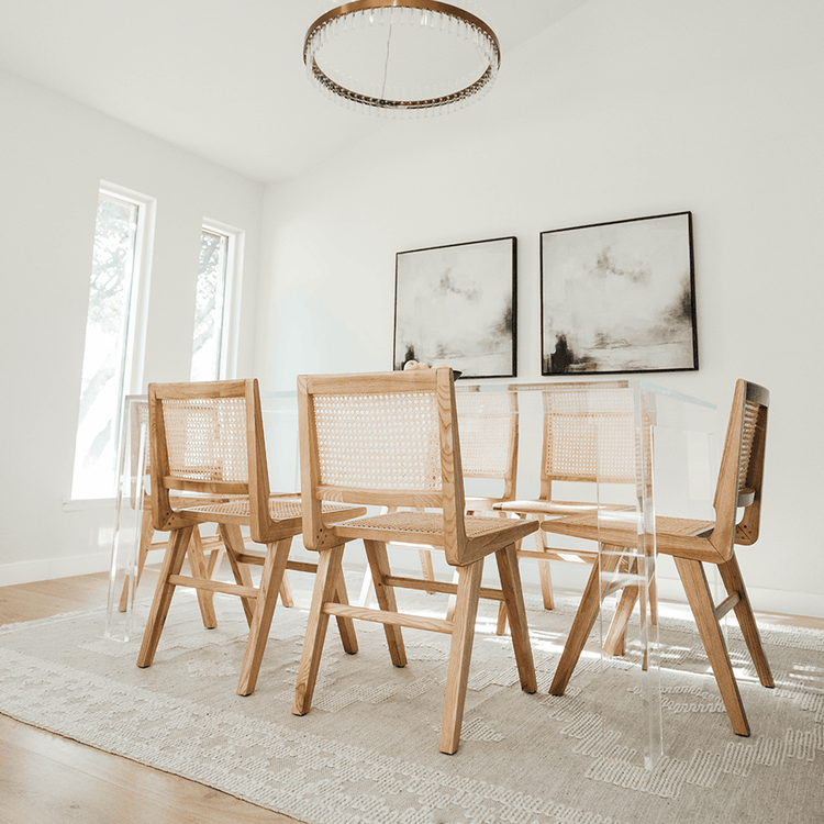 A clear acrylic lucite dining table decorated with a bowl of fruit surrounded by light wood and wicker chairs for seating in a brightly lit dining room.