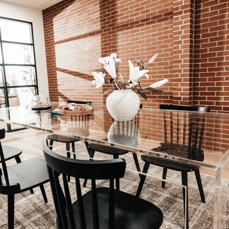 A clear acrylic lucite dining table decorated with snacks and a vase of flowers surrounded with black chairs.