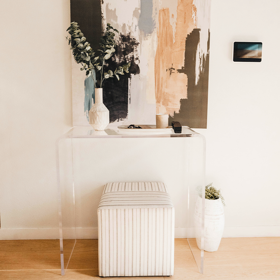 Clear acrylic waterfall edge entry or hallway table displaying a canvas painted picture, vase of flowers, decorative dish, and candle with an ottoman underneath against a wall in an entryway.