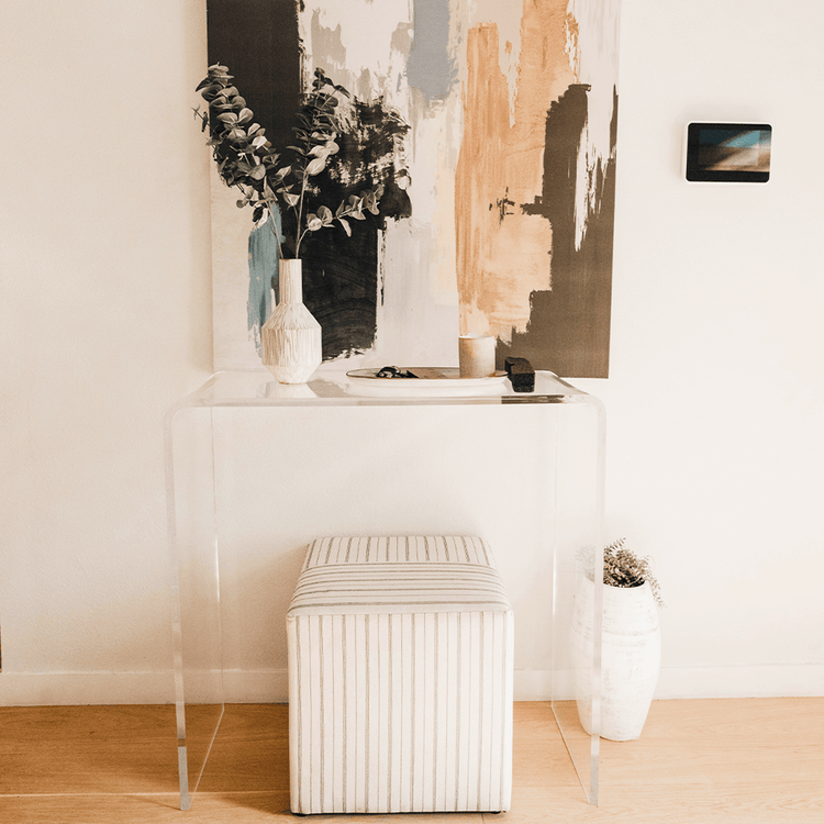 Clear acrylic waterfall edge entry or hallway table displaying a canvas painted picture, vase of flowers, decorative dish, and candle with an ottoman underneath against a wall in an entryway.