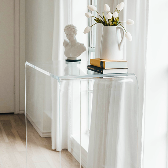 A clear acrylic waterfall console table decorated with a vase of tulips, books, and a bust statue placed in a brightly lit hallway in front of a window with white curtains.