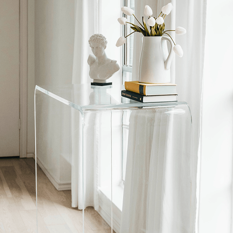 A clear acrylic waterfall console table decorated with a vase of tulips, books, and a bust statue placed in a brightly lit hallway in front of a window with white curtains.