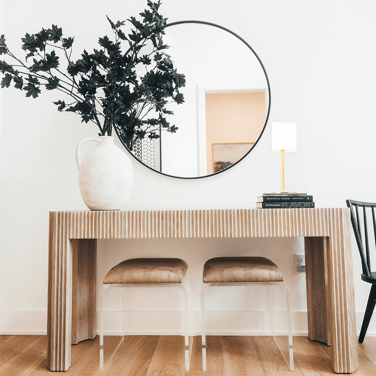 2 modern ottoman chairs with clear acrylic legs and brown leather seat upholstery, placed underneath an entryway table decorated with a vase of leaved branches, books with a lamp on top, and a wall mirror.