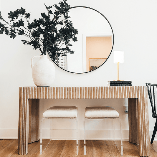 2 modern ottoman chairs with clear acrylic legs and white seat upholstery, placed underneath an entryway table decorated with a vase of leaved branches, books with a lamp on top, and a wall mirror.