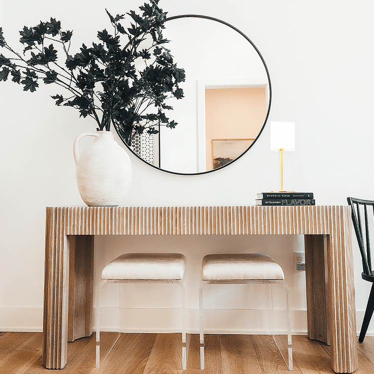 2 modern ottoman chairs with clear acrylic legs and white seat upholstery, placed underneath an entryway table decorated with a vase of leaved branches, books with a lamp on top, and a wall mirror.