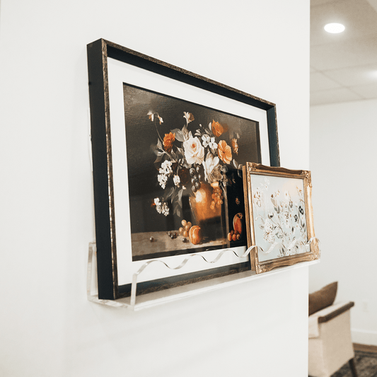 Framed floral artwork on a white wall being held by a clear acrylic floating bookshelf with a wavy edge.