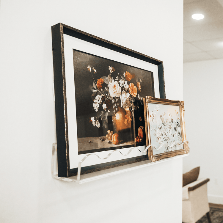 Framed floral artwork on a white wall being held by a clear acrylic floating bookshelf with a wavy edge.