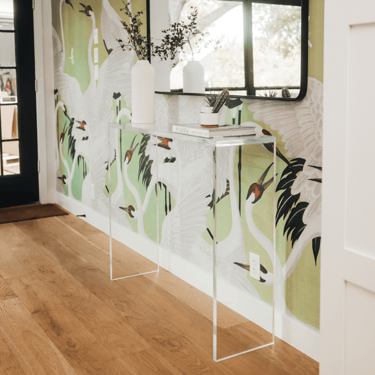 A clear acrylic console table decorated with plants and books placed against a wall with floral wallpaper.