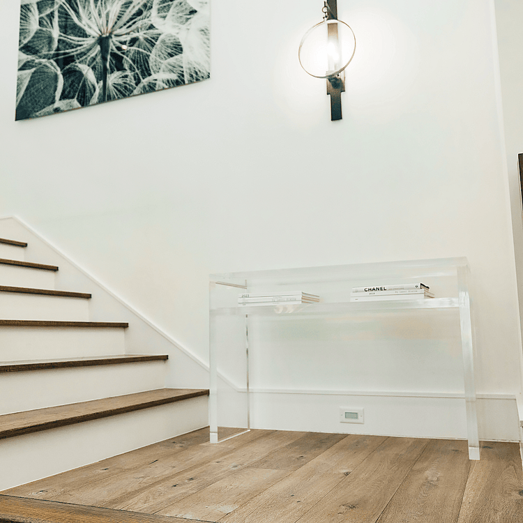 A clear acrylic console table with books on the interior shelf placed on a landing of a wooden staircase.