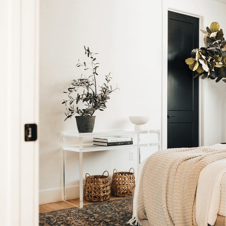 A clear acrylic lucite slab console table with a rectangular top and 1 interior shelf, placed in a bedroom with a potted plant and decorative items on top and baskets underneath.
