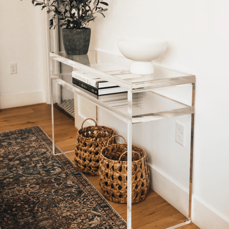 A clear acrylic lucite slab console table with a rectangular top and 1 interior shelf, placed in a bedroom with a potted plant and decorative items on top and baskets underneath.