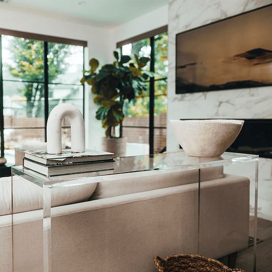 A modern clear acrylic console table with a slab style reflective surface, placed in a room with a beige sofa and decorative items on top.