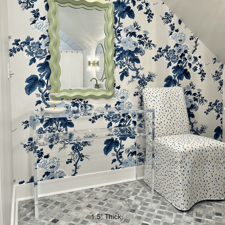 A clear acrylic console table with 1 interior shelf decorated with a jewelry dish placed against a wall with floral wallpaper and a mirror. An upholstered chair is placed beside.