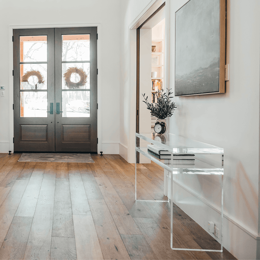 A clear acrylic console table with 1 interior shelf decorated with a plant and a clock placed against a wall with framed artwork in the entryway of a home.