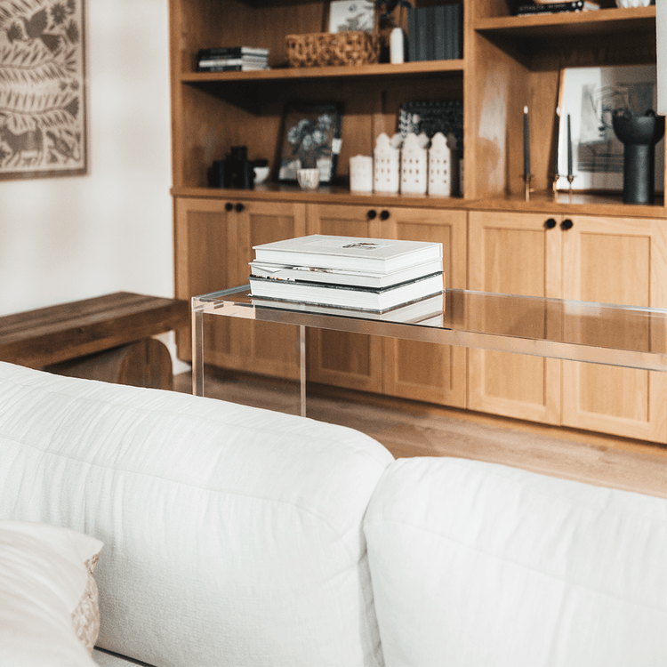 A modern clear acrylic console table with a slab style reflective surface, placed in a room with a beige sofa and books on top.