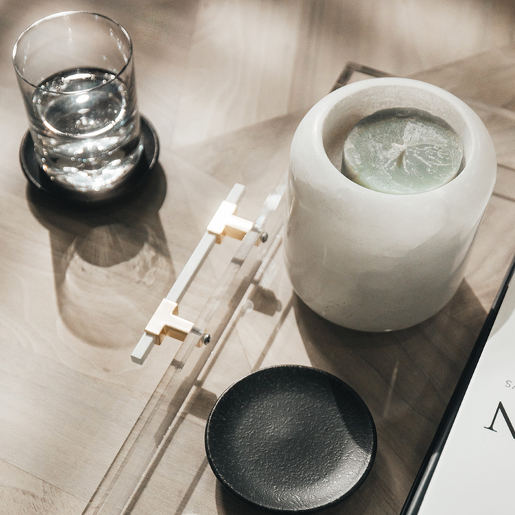 A clear acrylic lucite serving tray with gold and white handles holding decor, a candle, a glass, and books placed on a wooden table.