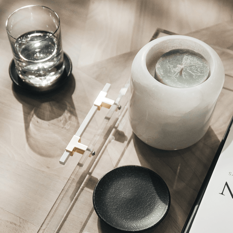 A clear acrylic lucite serving tray with gold and white handles holding decor, a candle, a glass, and books placed on a wooden table.