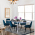 Clear acrylic slab style dining room table displaying centerpieces of flower vases, wine glasses, and place settings surrounded by chairs on an area rug in a dining room.
