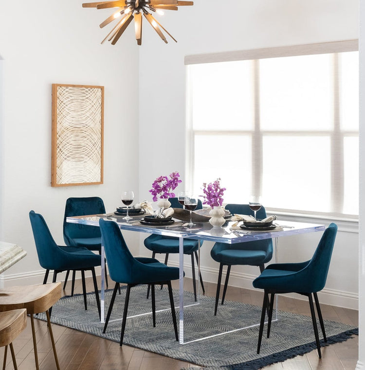 Clear acrylic slab style dining room table displaying centerpieces of flower vases, wine glasses, and place settings surrounded by chairs on an area rug in a dining room.