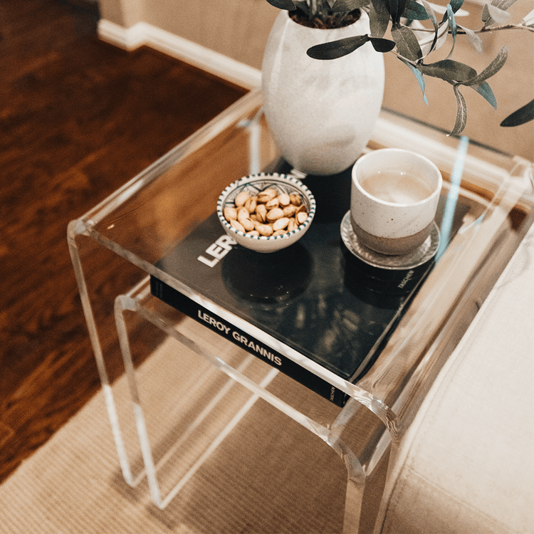 2 clear acrylic waterfall edge nesting end tables with book, vase, and decor next to a couch in a living room.
