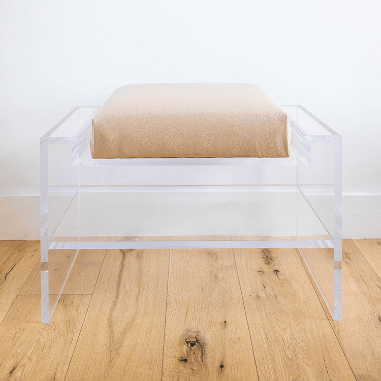 Catalog sample of a clear acrylic bench with a doe colored cushion and storage shelf underneath on a wooden floor.