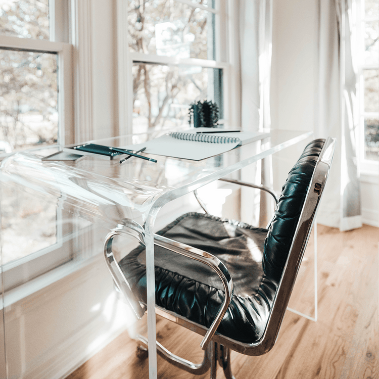 A closeup of the waterfall edge on a clear acrylic desk decorated with various office supplies and chair pushed underneath facing windows.