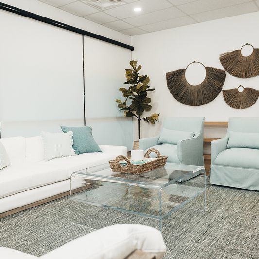 A clear acrylic waterfall edge coffee table in a living room setting in front of a couch featuring 1 interior shelf decorated with a basket and candle. 