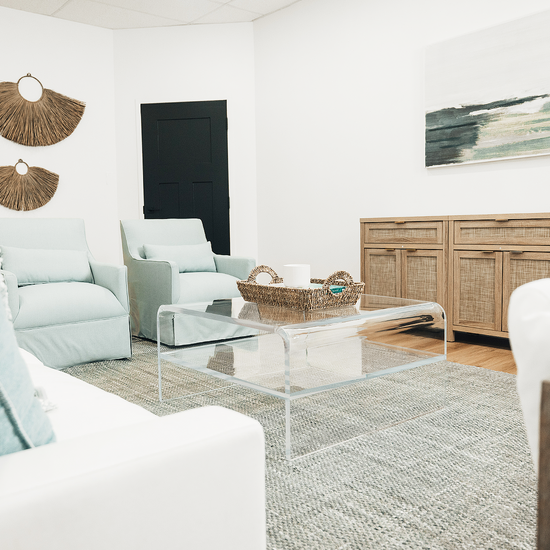 A clear acrylic waterfall edge coffee table in a living room setting in front of a couch featuring 1 interior shelf decorated with a basket and candle. 