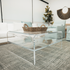 A clear acrylic square coffee table with a waterfall edge design and 1 interior shelf, placed in a living room setting with a white sofa, light blue easy chairs and decorative items.