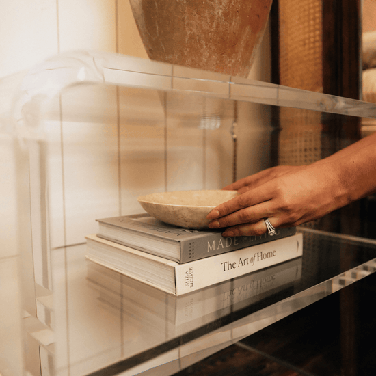 A person placing a decorative bowl on the interior shelf of a clear acrylic side table placed next to a bed.