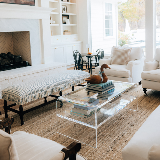A living room setting in front of a fireplace and bench featuring a clear acrylic waterfall edge coffee table placed on a rug.