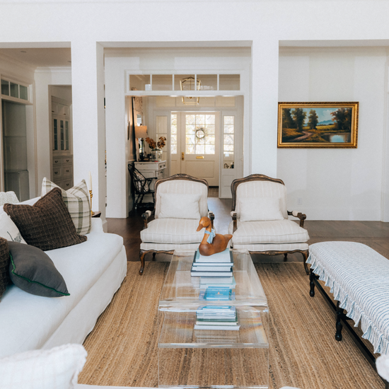 Living room with beige sofa, armchairs and a clear acrylic coffee table with 1 interior shelf displaying decorative items and books.