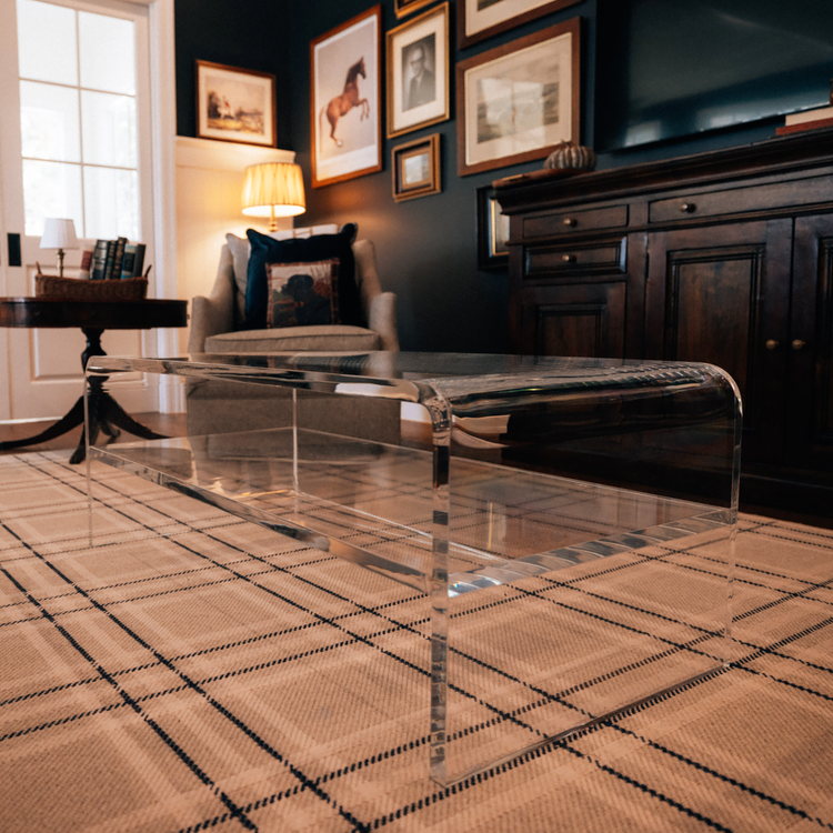 Living room with an upholstered armchair, credenza and a clear acrylic waterfall edge coffee table placed on a plaid rug. 