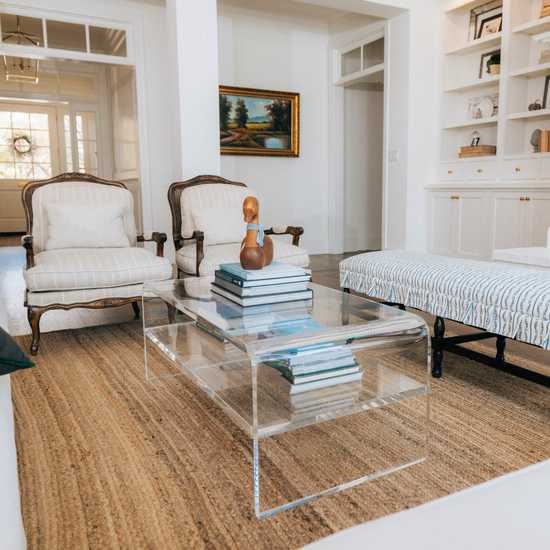 Living room with beige sofa, armchairs and a clear acrylic coffee table with 1 interior shelf displaying decorative items and books.