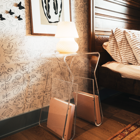 An acrylic lucite side table featuring legs that curve up into the inside of the table to double as a book or magazine rack displaying a lamp sitting beside a bed doubling as a nightstand.