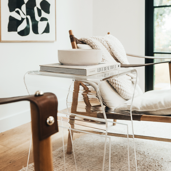 An acrylic lucite side table featuring legs that curve up into the inside of the table to double as a book or magazine rack sitting between 2 chairs in a living room.