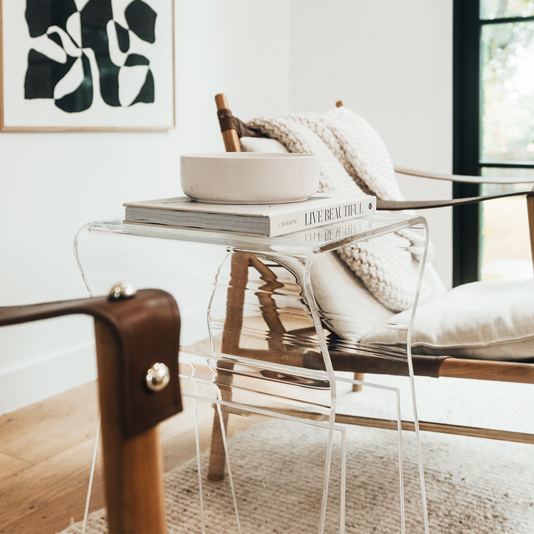 An acrylic lucite side table featuring legs that curve up into the inside of the table to double as a book or magazine rack sitting between 2 chairs in a living room.
