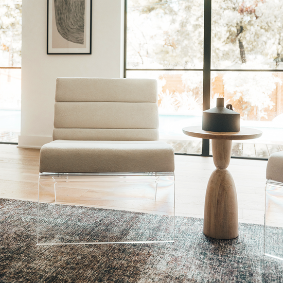 A clear acrylic lucite lounge chair with a white fabric seat and backrest, placed in a living room setting in front of large picture windows.