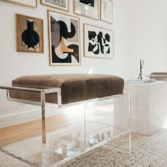 Closeup view of 1 of the two clear acrylic benches with brown cushions and storage shelf underneath, placed as extra seating in a modern living room with abstract art on the walls.