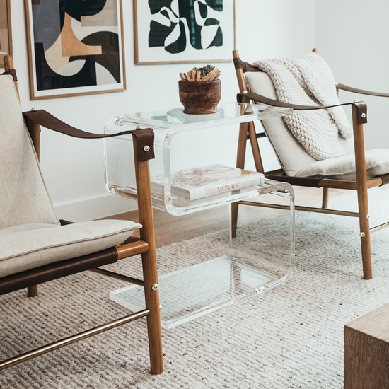 A clear acrylic side table shaped like the number 5 placed between 2 chairs in a living room.